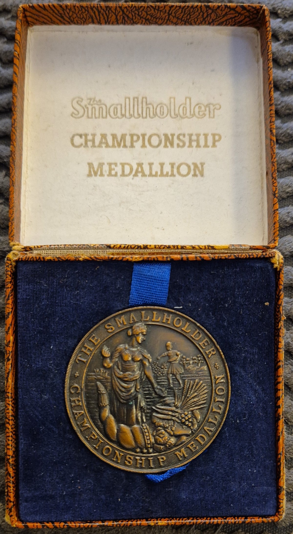 A round bronze-colored commemorative medallion displayed inside an open presentation box with a blue velvet insert and gold-toned trim. The inside lid reads 'Smallholder CHAMPIONSHIP MEDALLION.